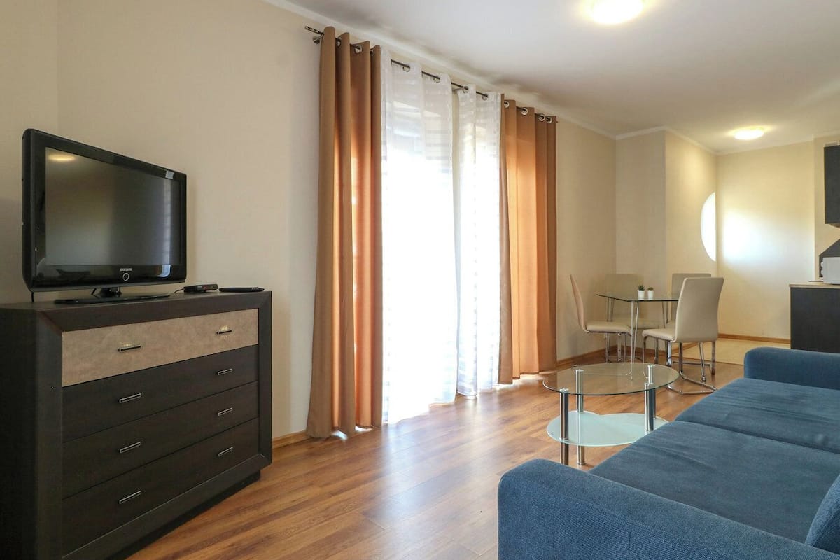 Un salon moderne avec un téléviseur à écran plat, une commode en bois foncé, un canapé bleu, une table à manger en verre avec des chaises, et une grande fenêtre avec des rideaux.