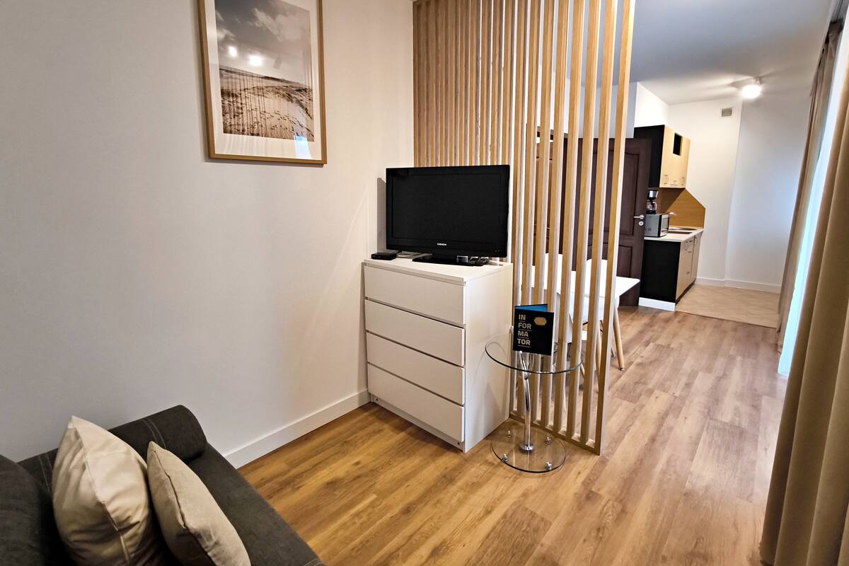 Un salon moderne avec un téléviseur à écran plat posé sur un meuble blanc, une table basse en verre, un plancher en bois et un coin cuisine visible à travers une cloison.
