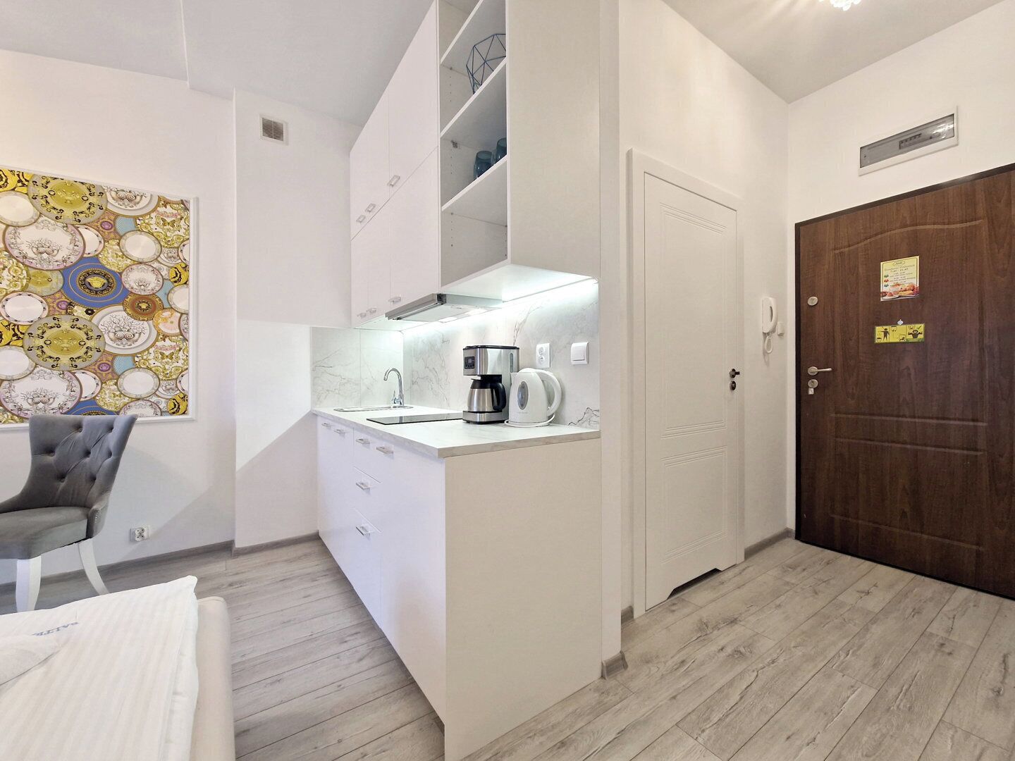 A compact kitchen with a built-in stove, a coffee maker, and a sink. There is a door with a sign on it, and a wall-mounted light fixture.