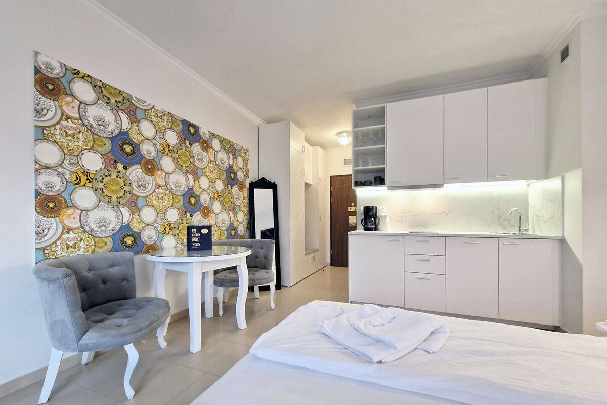 Une chambre d’hôtel moderne comprenant un lit, un fauteuil gris, une petite table ronde, une machine à café et un coin cuisine avec des placards blancs et un plan de travail en marbre.