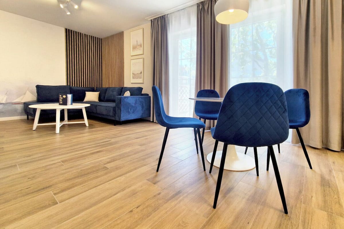 Un salon moderne avec un canapé, deux fauteuils bleus, une petite table blanche et une grande fenêtre avec des rideaux.