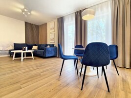 Ein modernes Wohnzimmer mit einer Couch, zwei blauen Sesseln, einem kleinen weißen Tisch und einem großen Fenster mit Vorhängen.