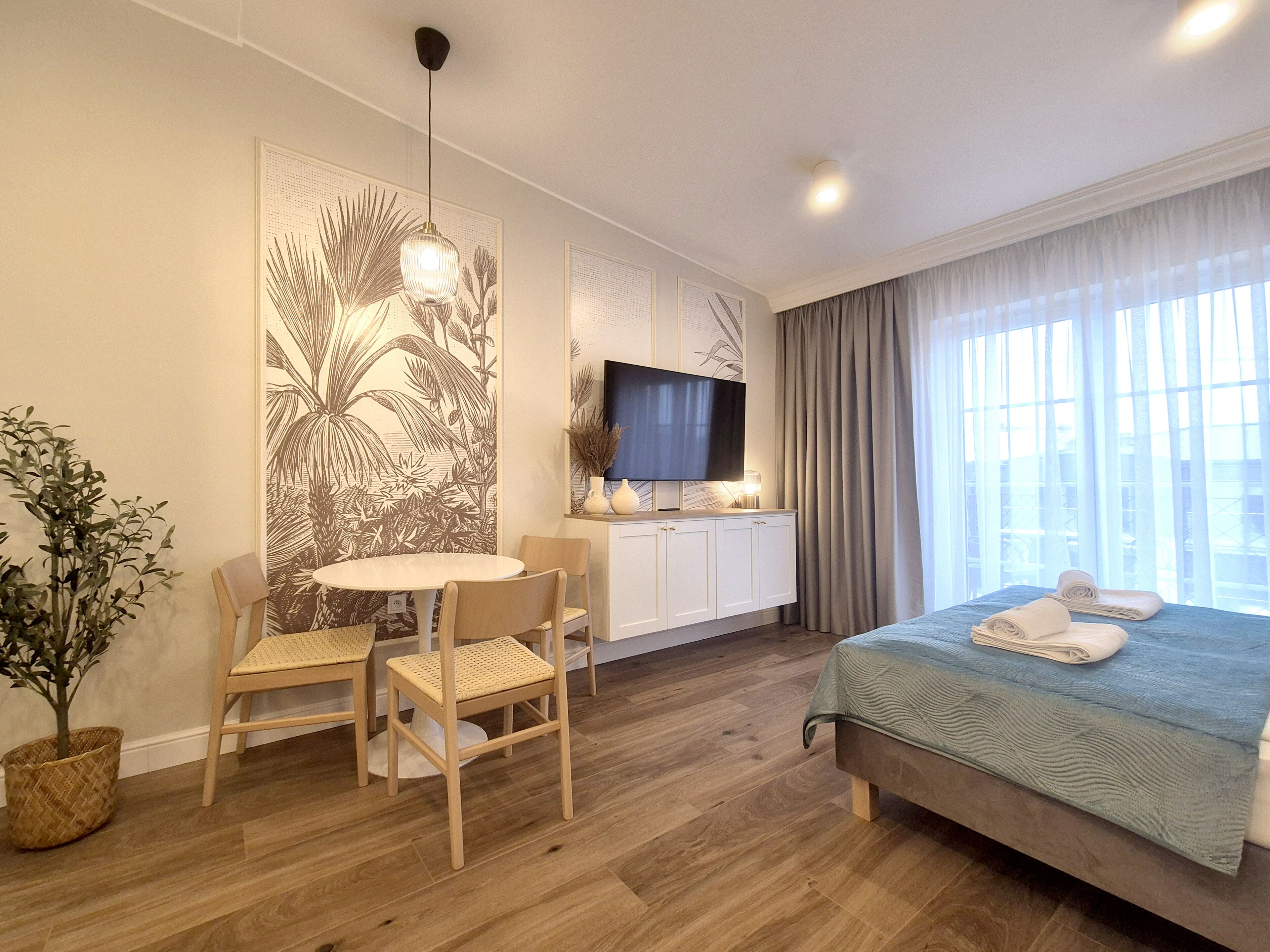 Une chambre d’hôtel comprenant un lit, une télévision, une table à manger avec des chaises, une grande œuvre d’art murale et une plante en pot.