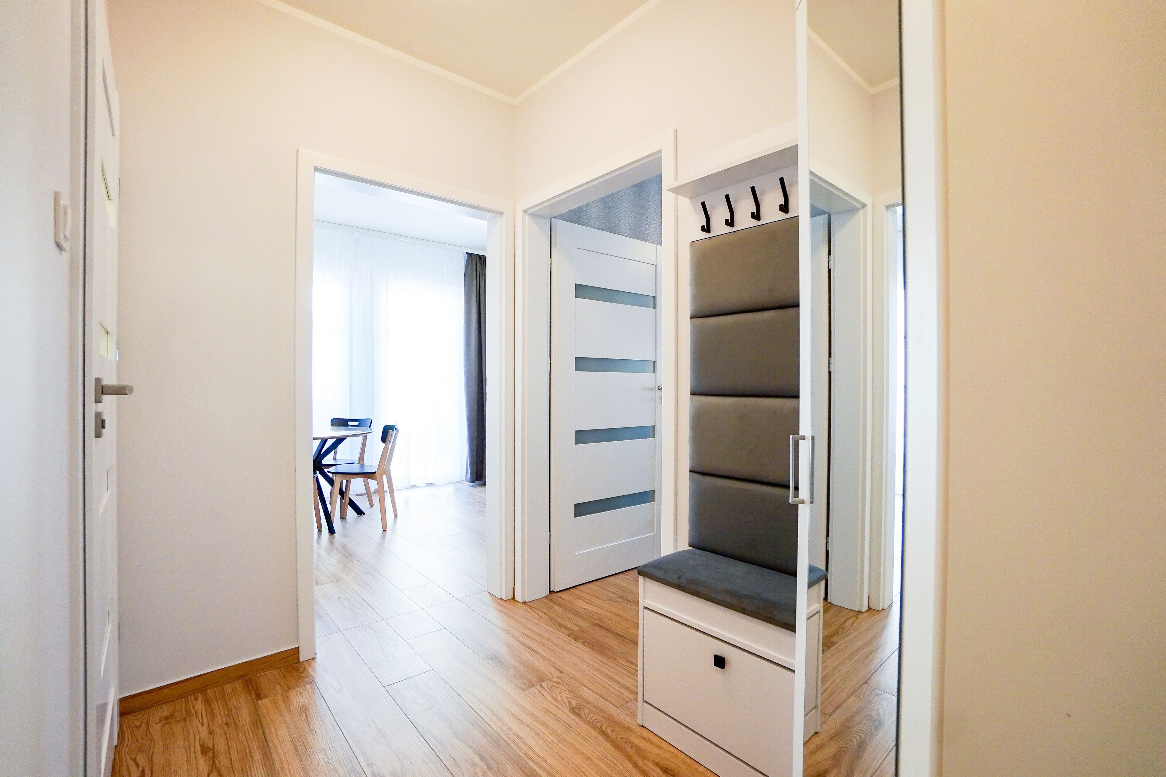 Un couloir avec un plancher en bois, une porte blanche et une pièce contenant un banc gris et une armoire blanche.