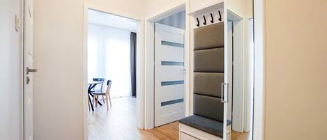 Un couloir avec un plancher en bois, une porte blanche et une pièce contenant un banc gris et une armoire blanche.