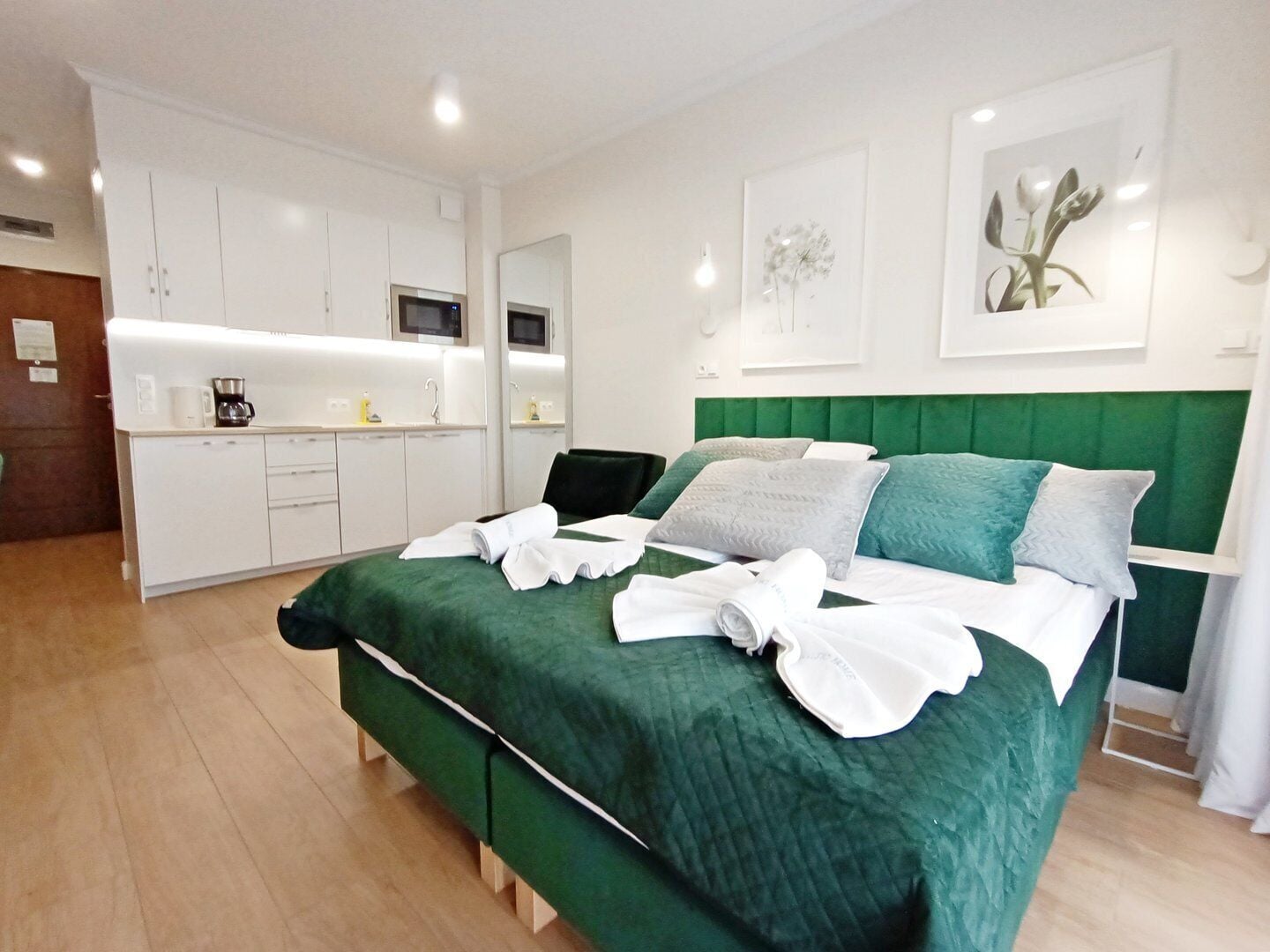 A modern hotel room with a green upholstered bed, white linens, a kitchenette, and a large window.