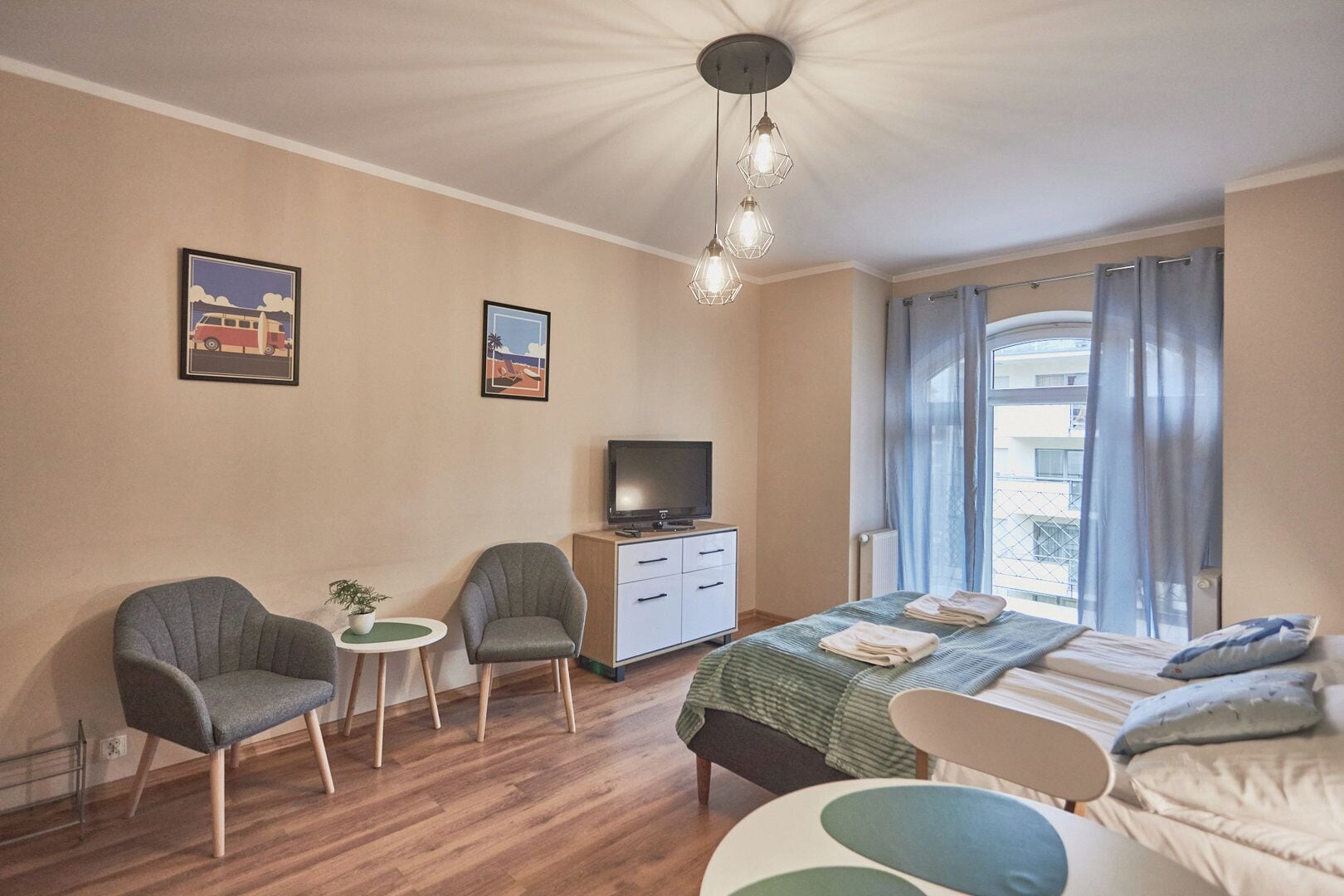 A hotel room with a bed, two armchairs, a small table with a plant, a TV, and a dresser.