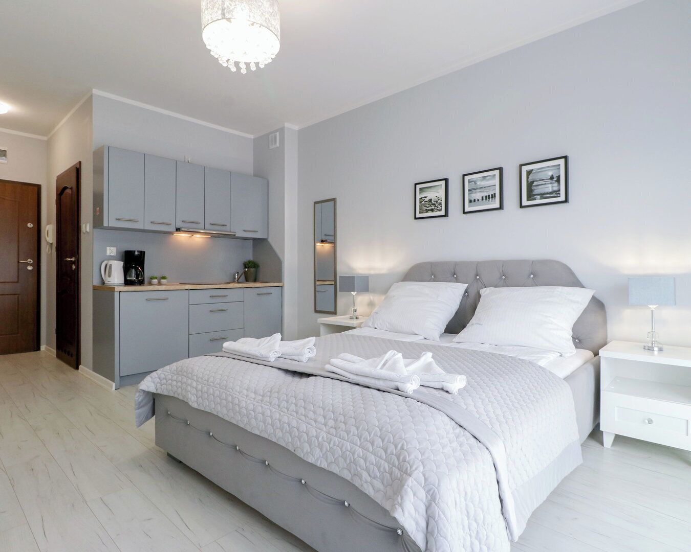 A modern bedroom with a large bed, a bedside table, a nightstand, a built-in kitchen area, and a mirror.