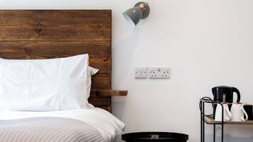 A neatly made bed with white linens, a wooden headboard, a wall-mounted lamp, a power outlet, and a small table with a kettle and mug.