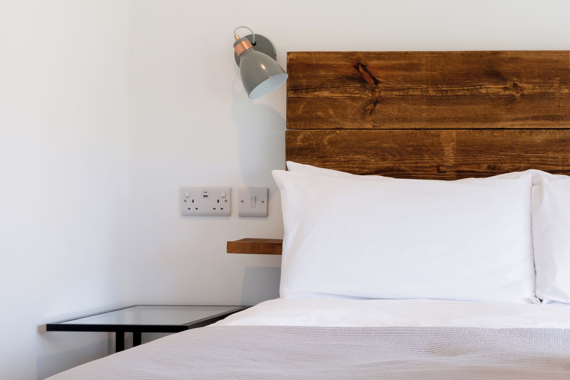 Un dormitorio con cabecera de madera, ropa de cama blanca, una mesita de noche y una lámpara de pared.