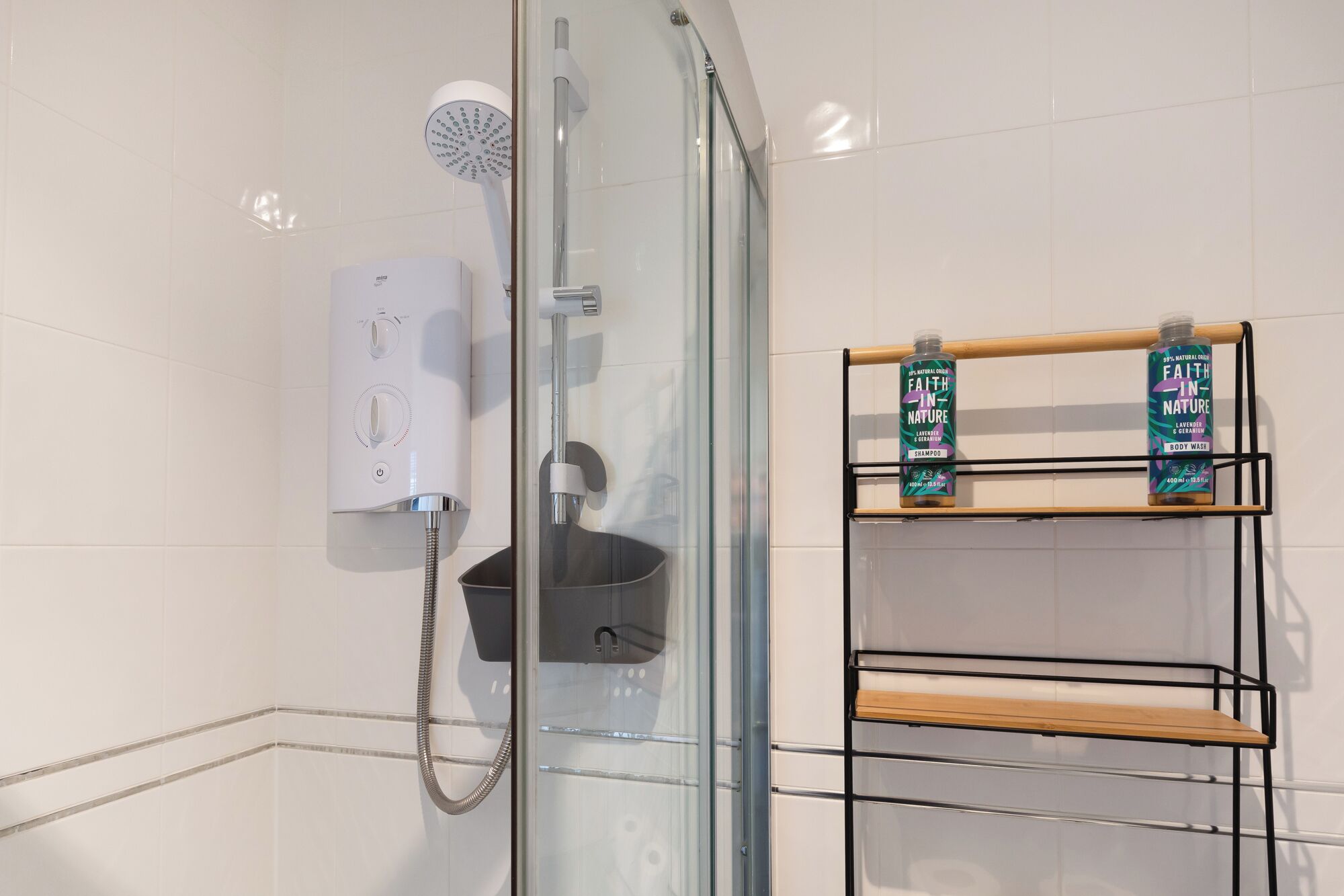 A modern bathroom with a glass-enclosed shower, a handheld showerhead, and a built-in water heater. There are two shelves with bottles of shampoo and body wash.