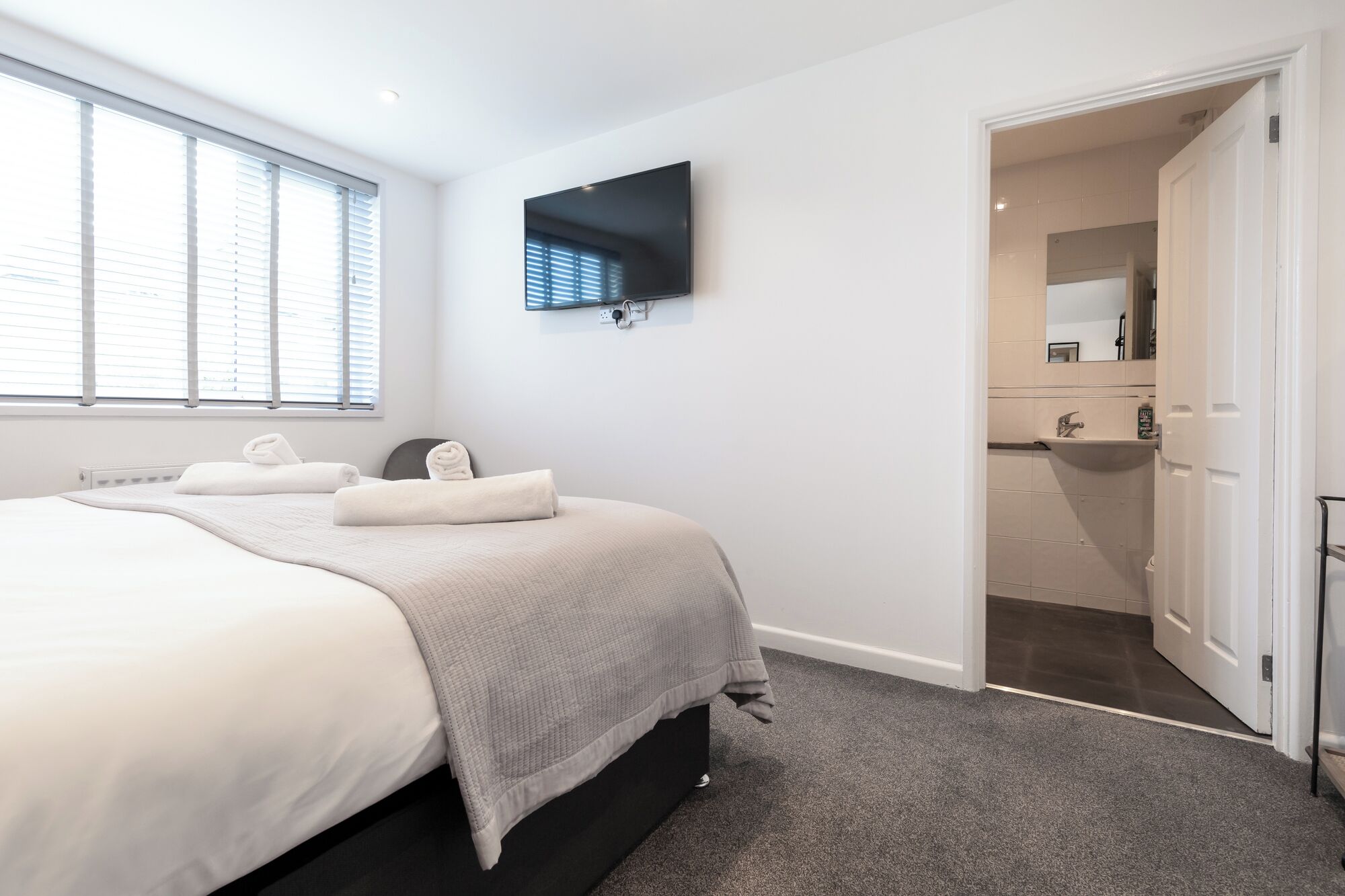 A modern bedroom with a large bed, a flat-screen TV mounted on the wall, and a bathroom visible through an open door.