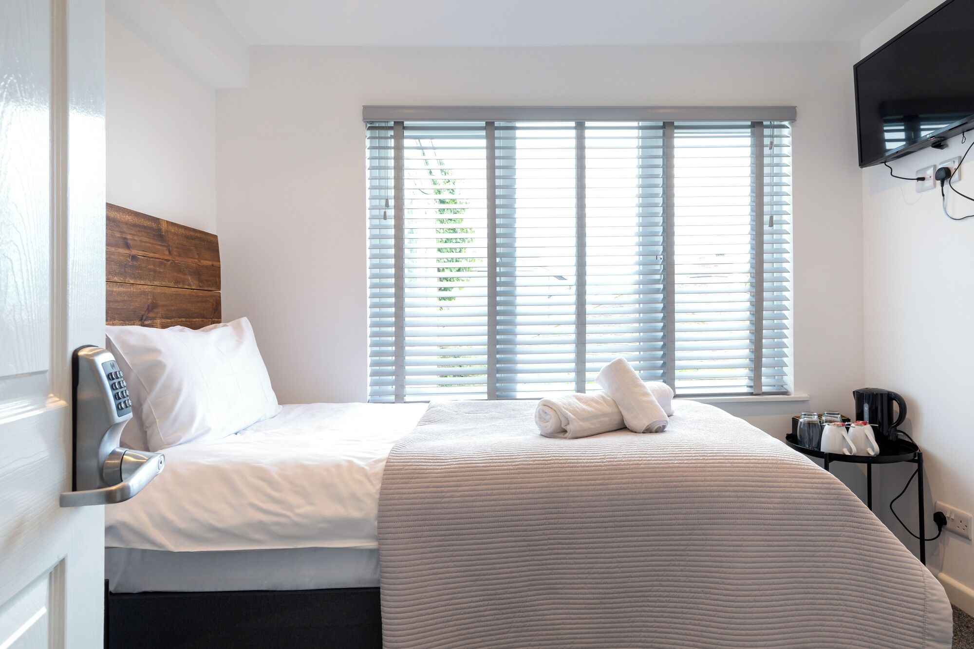 A hotel room with a bed, a window with blinds, a bedside table with a lamp, and a wall-mounted TV.