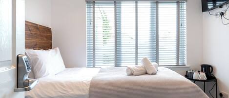 A hotel room with a bed, a window with blinds, a bedside table with a lamp, and a wall-mounted TV.