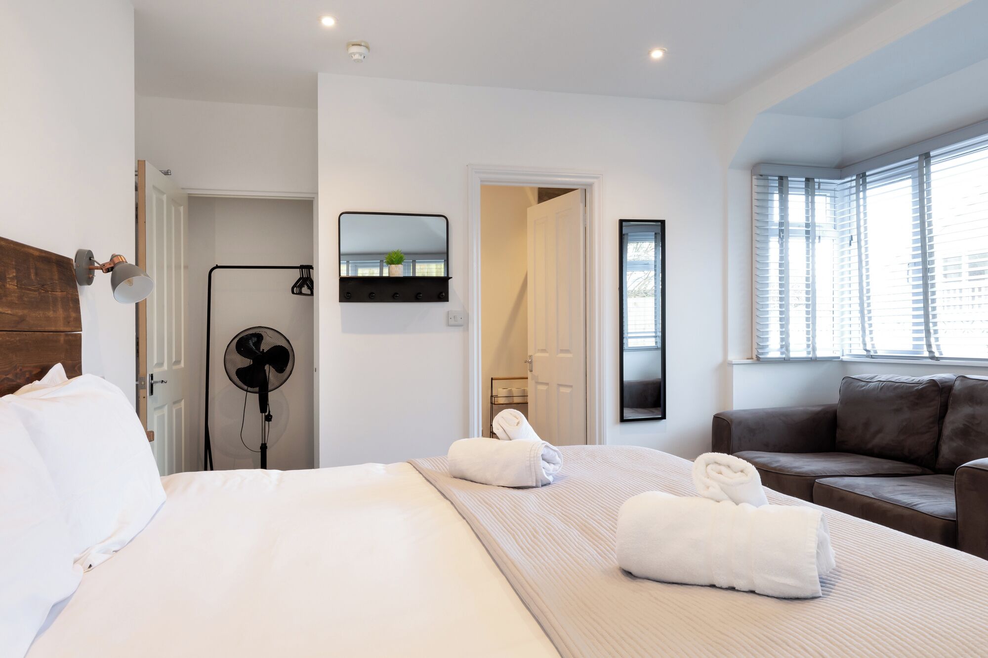 Habitación de hotel con una cama grande, una zona de estar con sofá, un ventilador, un espejo y una ventana con persianas.