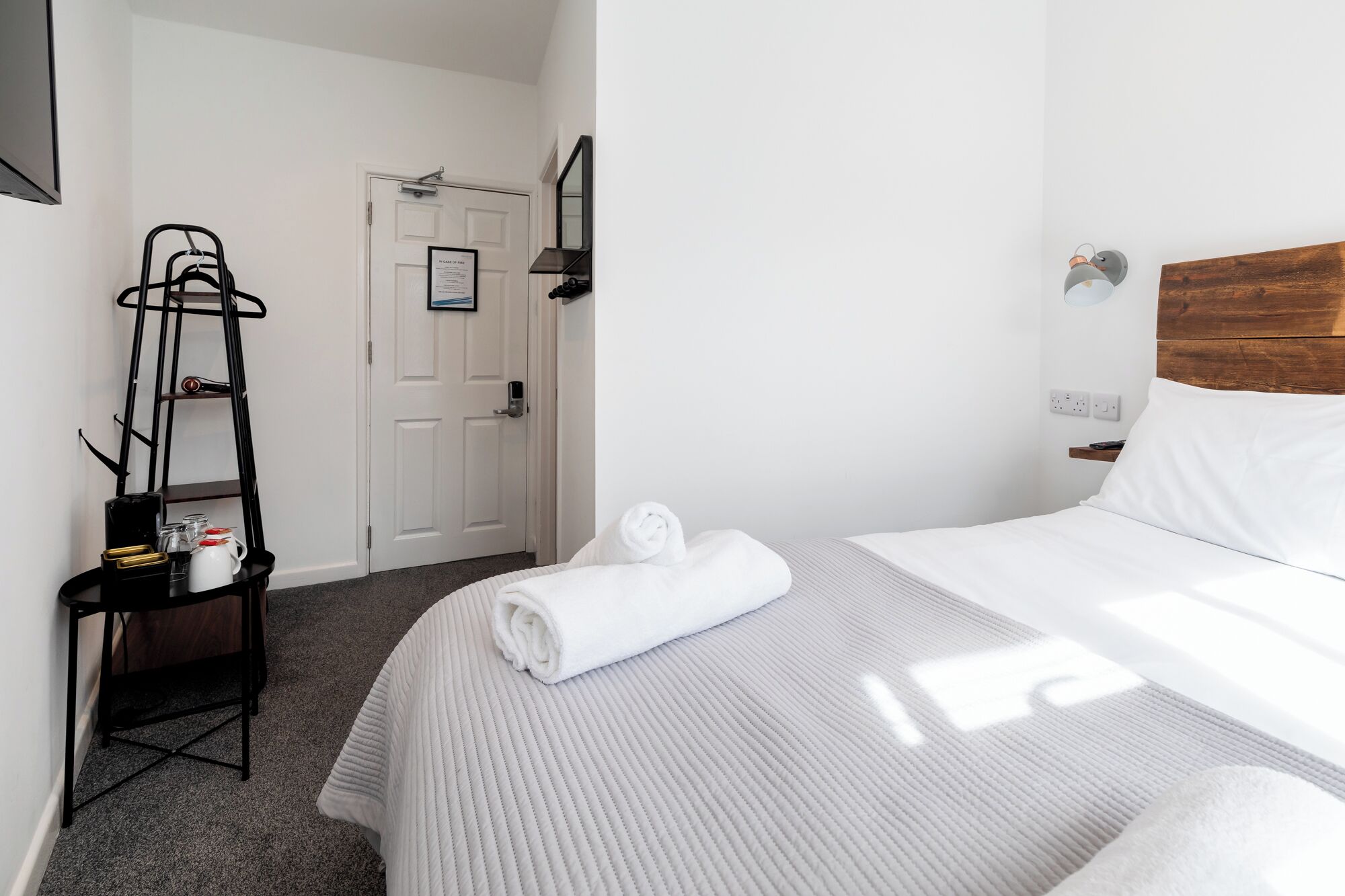 A hotel room with a bed, a towel, a shelf with toiletries, a door, and a wall-mounted light fixture.
