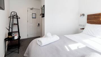 A hotel room with a bed, a towel, a shelf with toiletries, a door, and a wall-mounted light fixture.