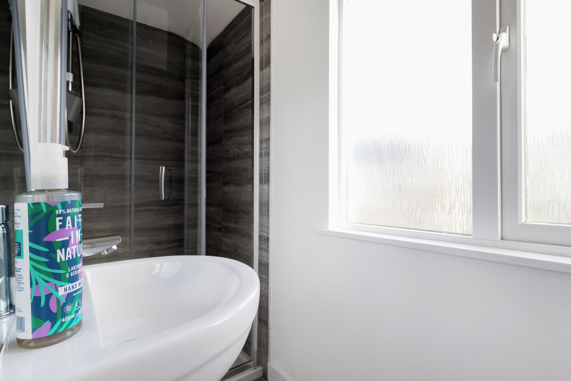 Un baño con un lavamanos blanco, una ducha con puerta de vidrio y una ventana con cristal esmerilado.