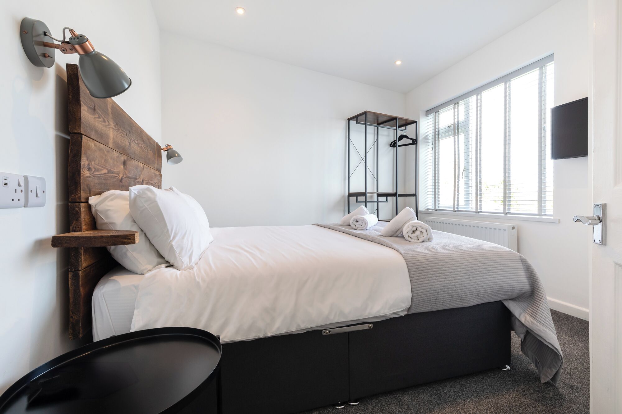 Un dormitorio moderno con cabecera de madera, cama con ropa de cama blanca, mesita de noche negra, lámpara de pared, espejo y ventana con persianas.