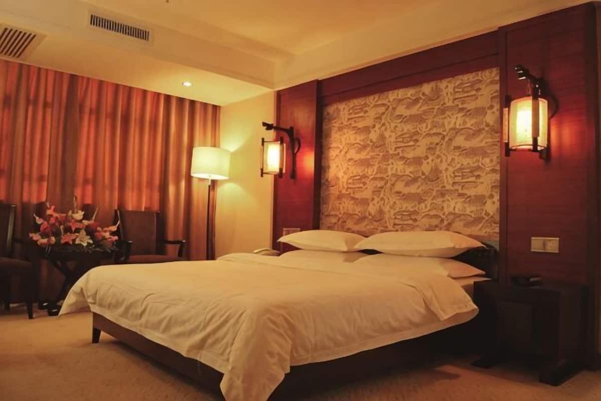 Une chambre d’hôtel avec un grand lit, deux lampes de chevet, une tête de lit en bois, une table de chevet et une chaise agrémentée d’un vase de fleurs.