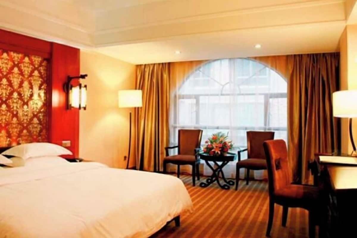 Une chambre d’hôtel avec un grand lit, deux chaises, une petite table ornée de fleurs et une fenêtre avec des rideaux.