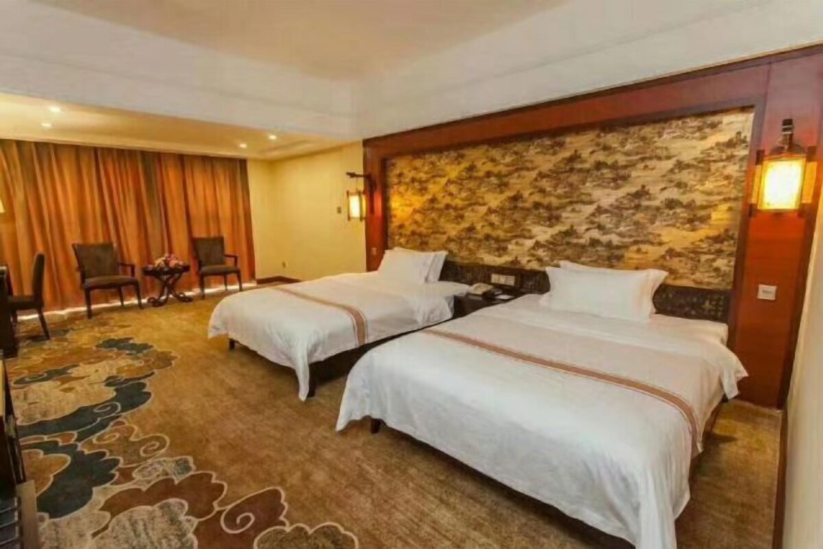 A hotel room with two beds, a carpeted floor, a headboard with a patterned design, and a lamp on the wall.