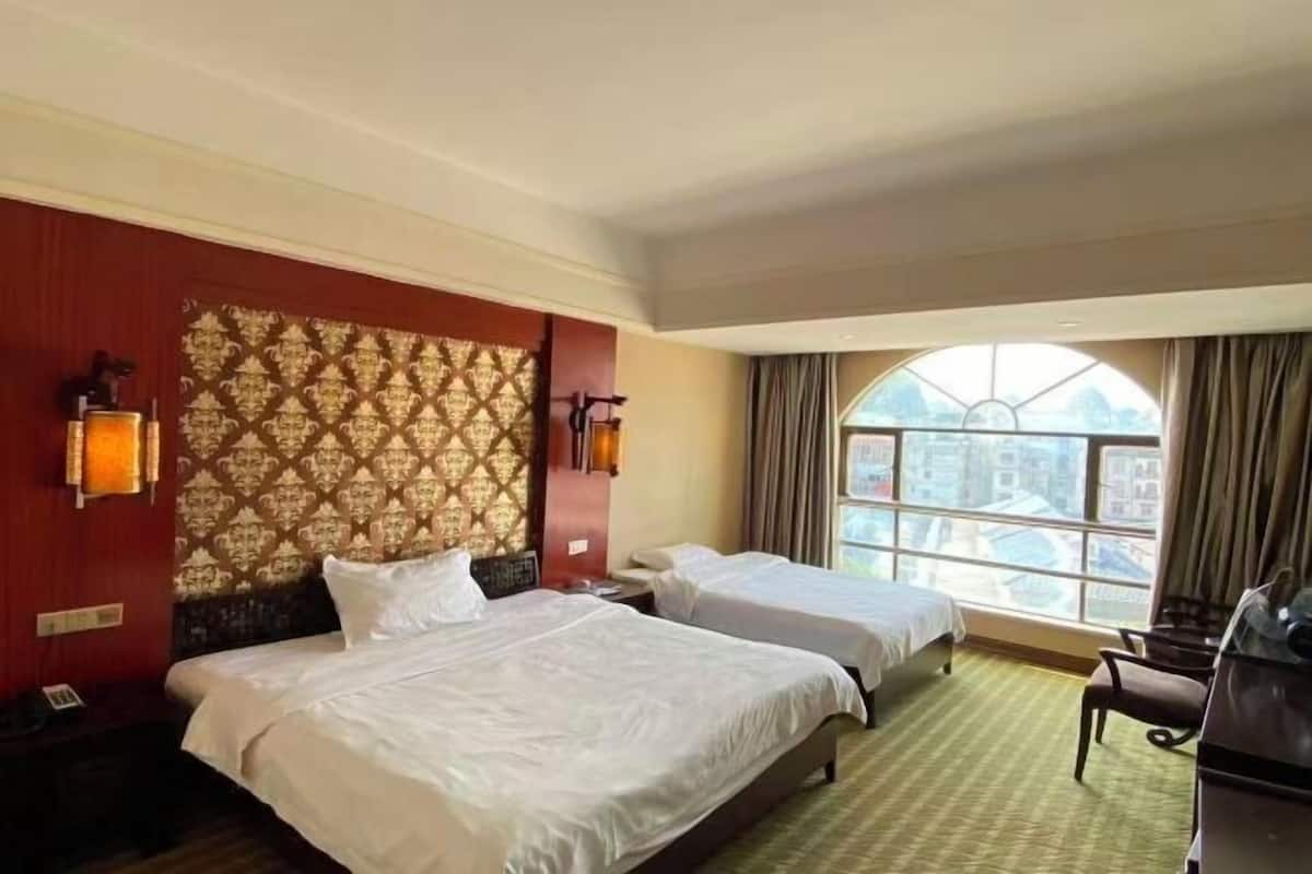 Une chambre d’hôtel avec deux lits, une grande fenêtre donnant sur la ville, un mur décoré d’un motif et un bureau avec une chaise.