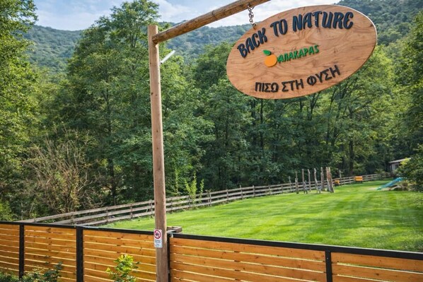 A wooden sign with 'BACK TO NATURE' and 'ALAKAPAS' text, a wooden fence, and a grassy area with trees in the background. - Scenic Mountain Getaway for Couples with Pool Access (Arakapas)
