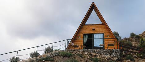 A wooden cabin with a triangular roof, a stone foundation, and a small porch with two chairs.