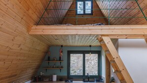 A loft-style room with a wooden sloped ceiling, a kitchen area with black cabinets, a wooden dining table, and a window with a view of trees. - Scenic Mountain Getaway for Couples with Pool Access (Arakapas)