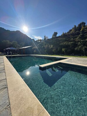 A clear swimming pool with a stone border, surrounded by a landscaped area with trees and a mountain in the background. - Scenic Mountain Getaway for Couples with Pool Access (Arakapas)
