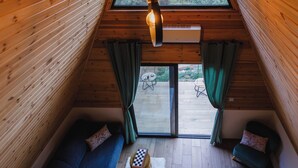 A cozy attic room with wooden walls, a sloped ceiling, a hanging light fixture, a blue sofa, a small table, and a view of greenery outside. - Scenic Mountain Getaway for Couples with Pool Access (Arakapas)