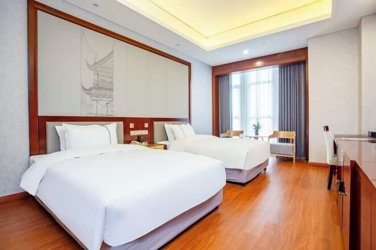 A hotel room with two beds, a wooden floor, a large window with curtains, and a desk with a chair.