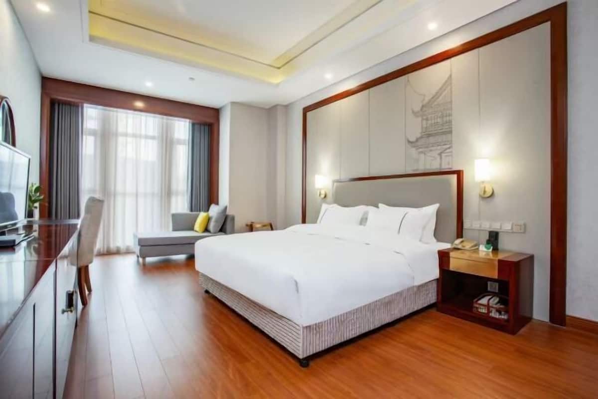 A modern hotel room with a large bed, a desk, a chair, and a nightstand with a lamp.