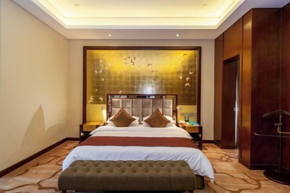 A hotel room with a large bed, a headboard with a decorative panel, a bedside table with a lamp, and a wooden wardrobe.