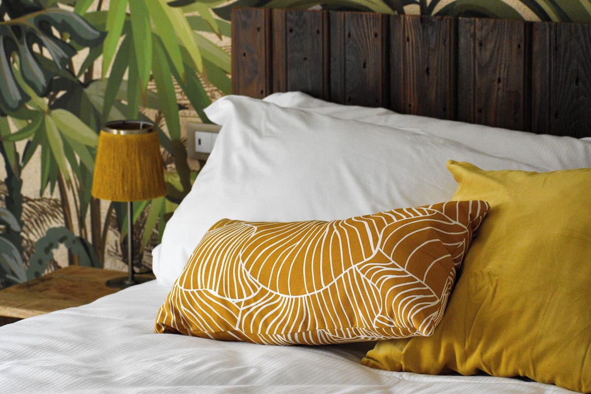 A bed with white and mustard-colored pillows, a wooden headboard, and a lamp on a wooden nightstand.