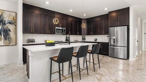 Una cocina moderna con armarios de madera oscura, una isla blanca con taburetes de bar, electrodomésticos de acero inoxidable y suelo de mármol.
