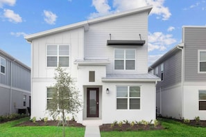A modern two-story residential building with a dark brown front door, white siding, and a small tree in the front yard.