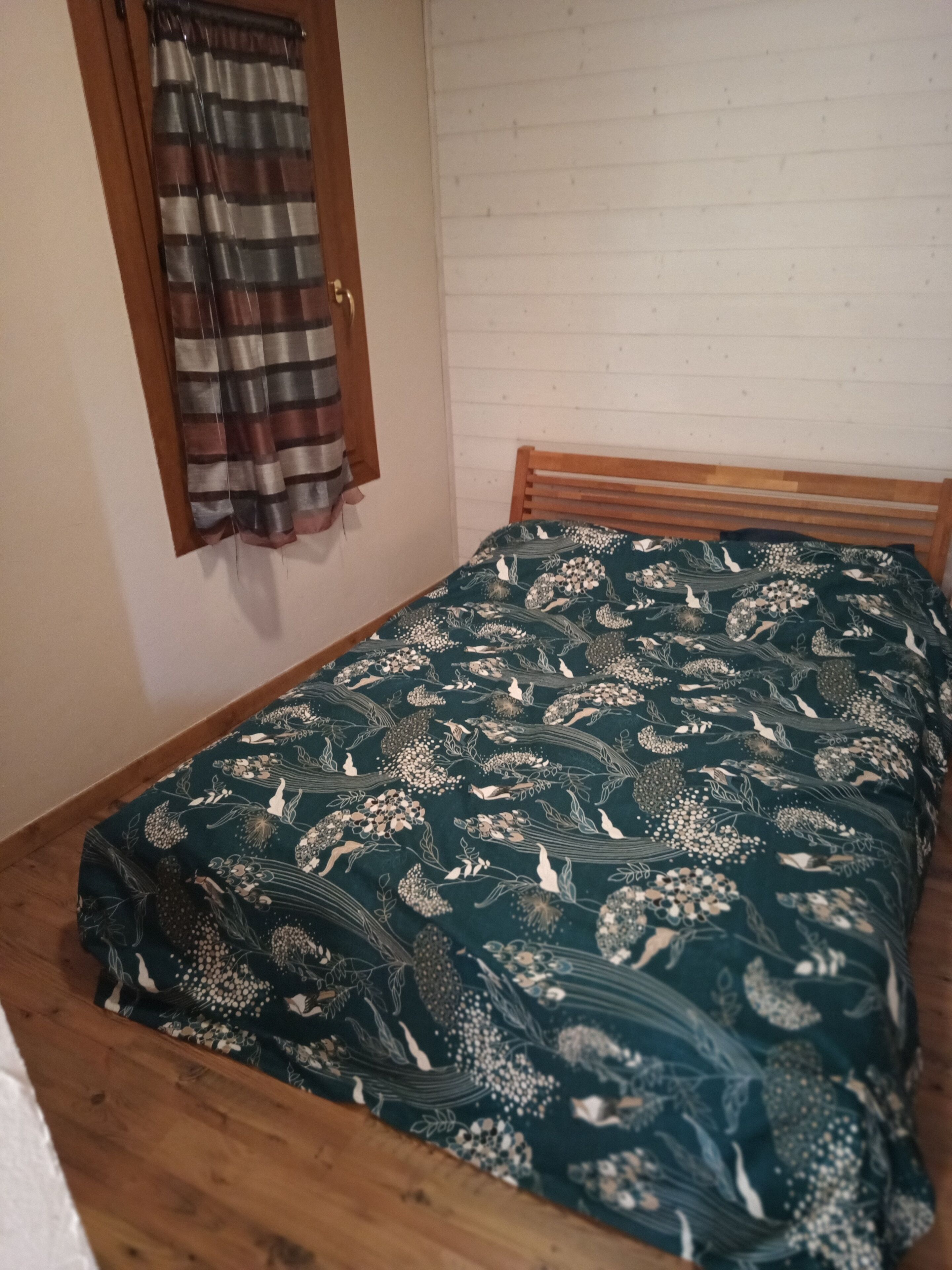 A small room with a wooden bed, a window with curtains, and a white brick wall.