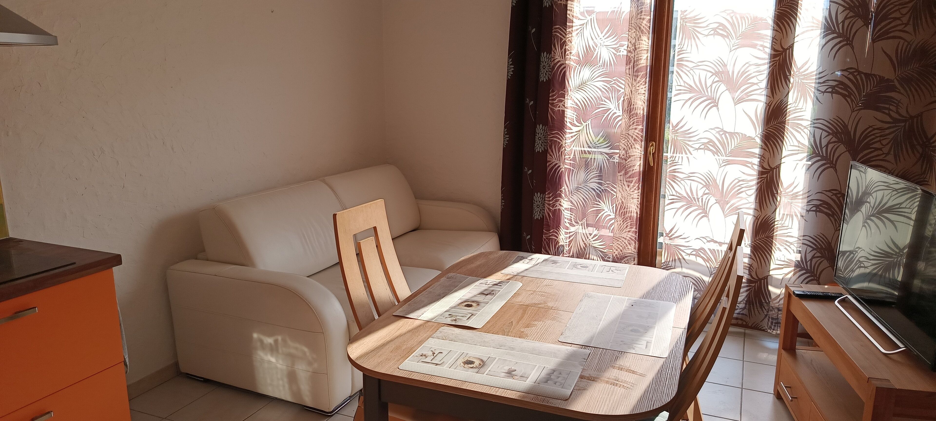 Een knus woongedeelte met een bank, een houten eettafel met placemats, een televisie op een houten dressoir, en een raam met geruite gordijnen.