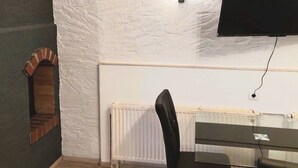Une pièce avec un plafond blanc à texture, une cheminée en briques, un radiateur blanc, une chaise noire et un téléviseur noir fixé au mur.