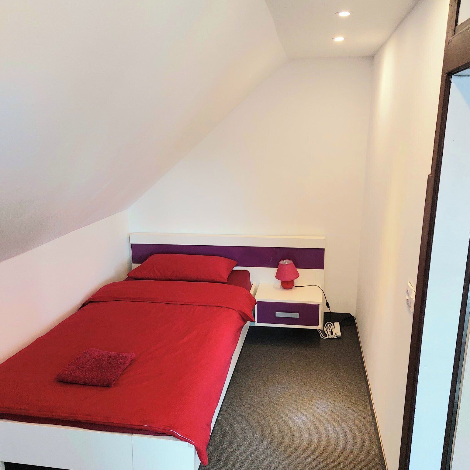 A small, neatly arranged bedroom with a bed, a bedside table with a lamp, and a drawer unit.