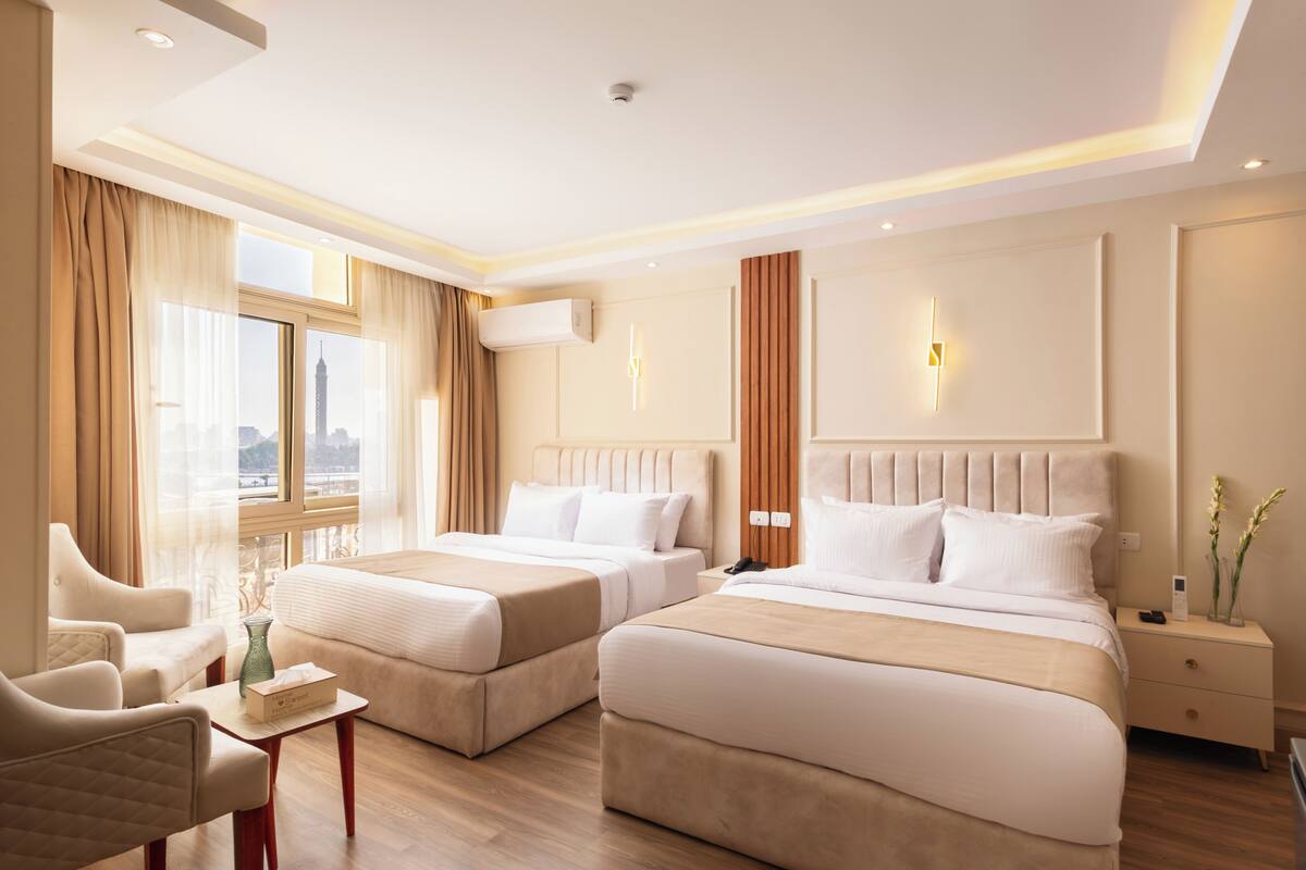 A hotel room with two beds, a sitting area with a chair, a small table, and a nightstand with a lamp.