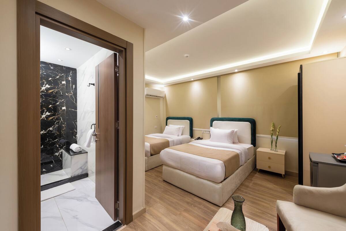 A modern hotel room with two beds, a wooden floor, a marble-tiled bathroom, and a small desk with a vase.
