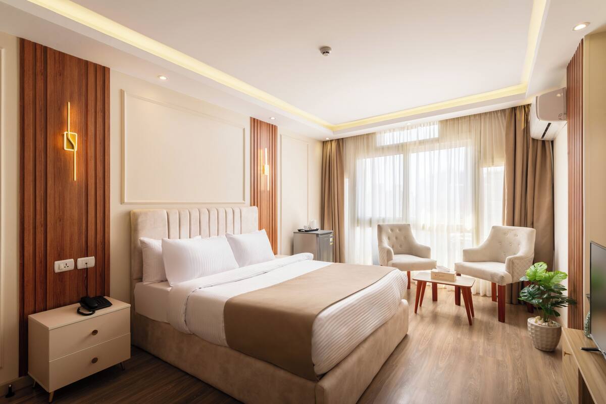 A modern hotel room with a large bed, bedside tables, a desk, two chairs, a television, and a potted plant.