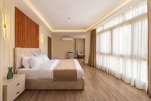 A modern hotel room with a large bed, wooden flooring, and large windows with sheer curtains.