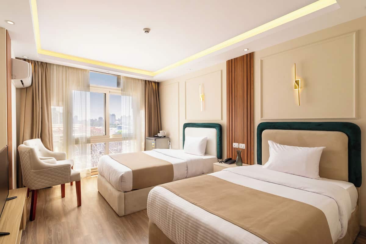 A hotel room with two beds, a sitting area with a chair, a desk, and a view of the city.