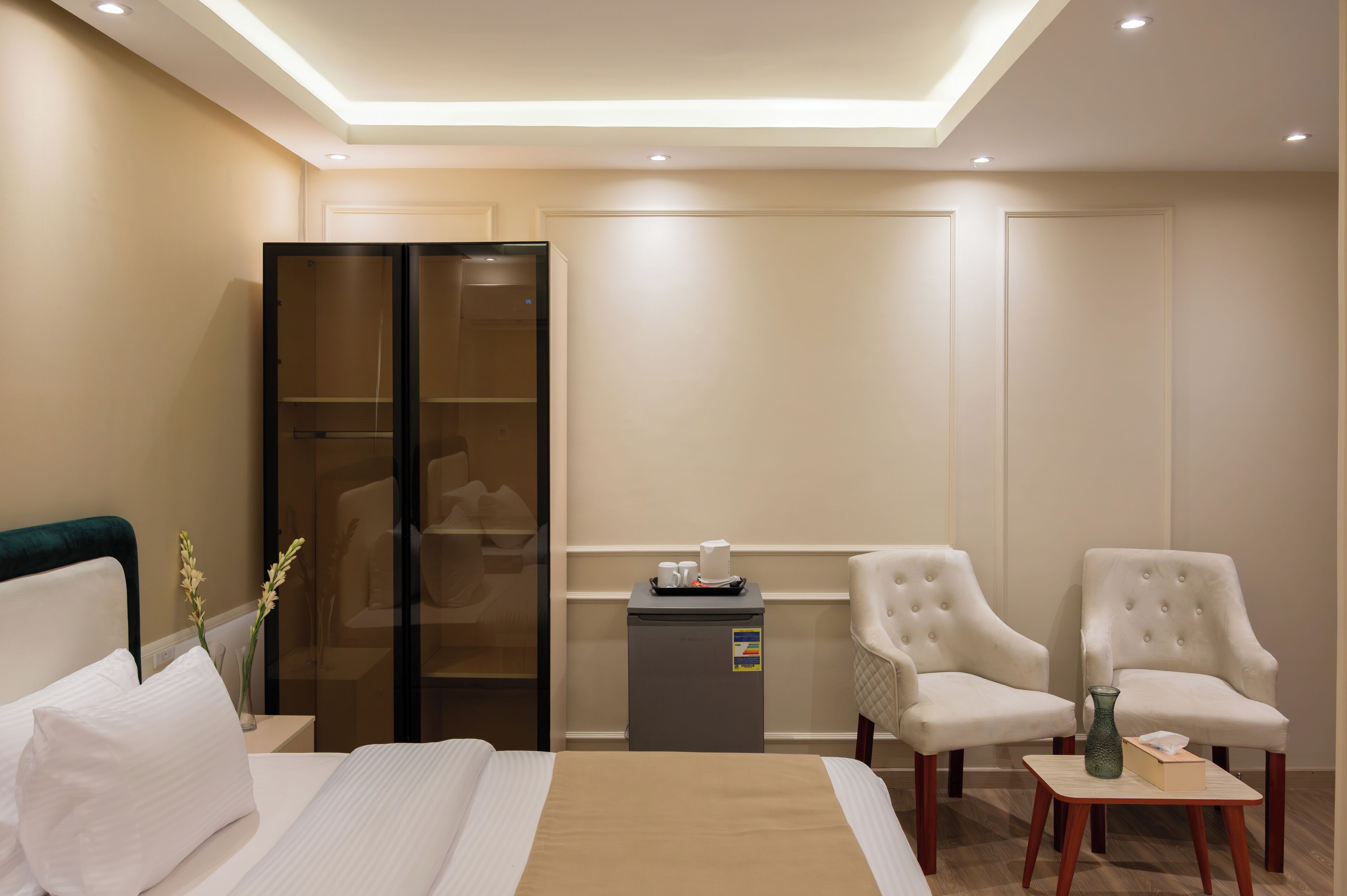 Ein Hotelzimmer mit einem Bett, einer Minibar, zwei Sesseln, einem kleinen Tisch mit einer Vase und einem gläsernen Schrank.