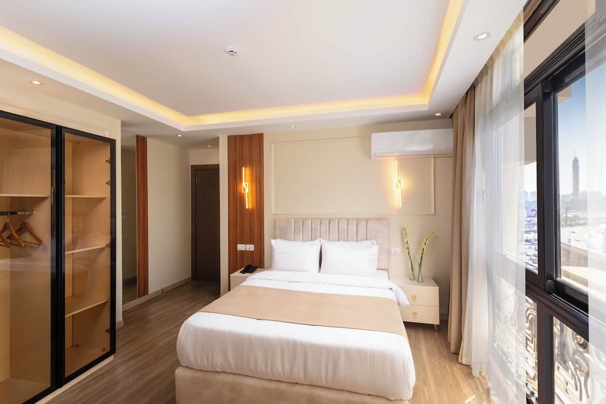 A modern hotel room with a large bed, a walk-in closet, a bedside table with a lamp, and a view of the city through large windows.