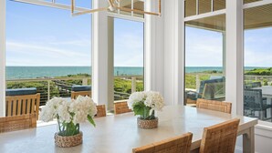 Un espace repas offrant une vue sur l’océan, de grandes fenêtres, des chaises en osier et une table décorée de fleurs blanches.
